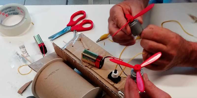 Foto evocativa per il Laboratorio STEMLAB dove si vedono mani impegnate nella costruzione di qualcosa, maneggiando forbici, cartone, piccole eliche e batterie.